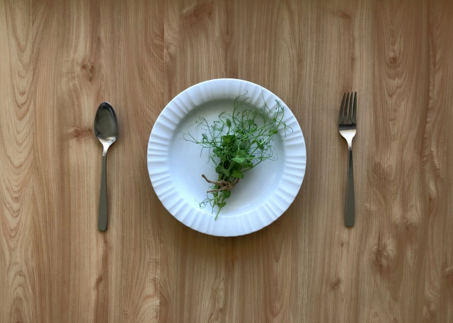 木目のテーブルの上にある白いお皿に、少量のスプラウト（芽野菜）だけが盛り付けられた様子。極端な食事制限や無理なダイエットを象徴するイメージ。