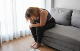 ビングのソファに座り、体重計を前にして頭を抱え、ダイエットの失敗や先延ばしに後悔し、落ち込む太り気味の女性。