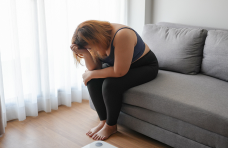 ビングのソファに座り、体重計を前にして頭を抱え、ダイエットの失敗や先延ばしに後悔し、落ち込む太り気味の女性。