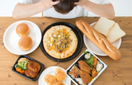 ピザ、パン、ハンバーガー、揚げ物弁当など、大量の高カロリー食品を前にして、食べ過ぎや食欲に悩んで頭を抱える女性。