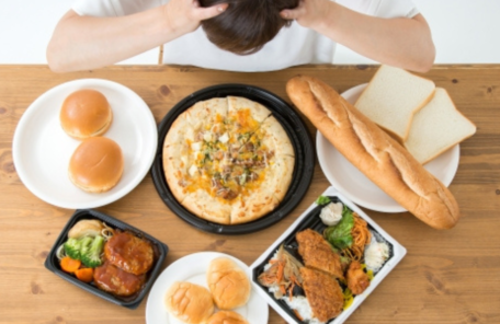 ピザ、パン、ハンバーガー、揚げ物弁当など、大量の高カロリー食品を前にして、食べ過ぎや食欲に悩んで頭を抱える女性。
