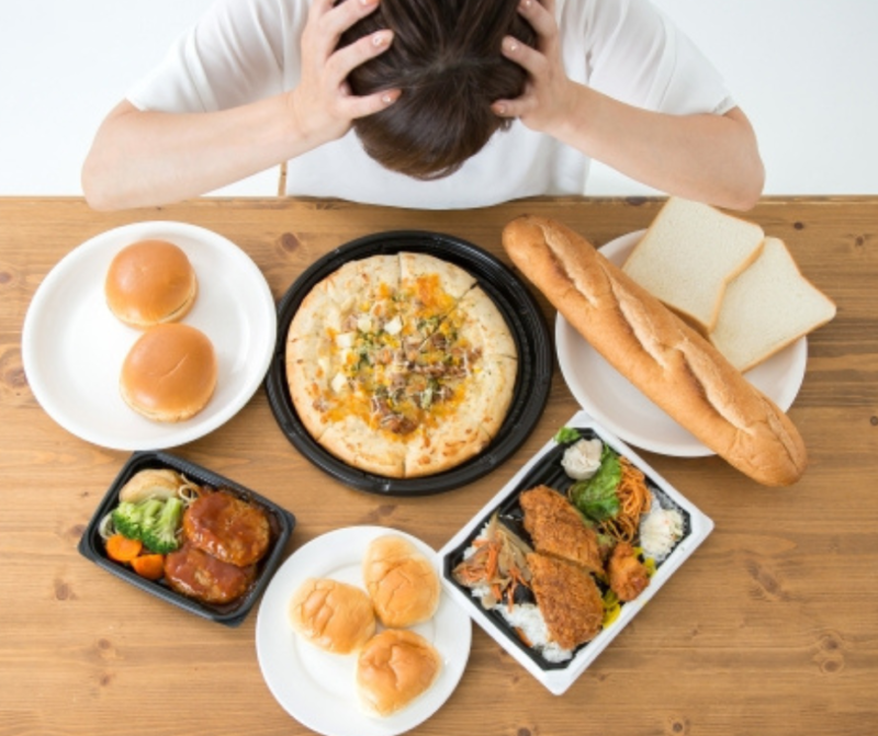ピザ、パン、ハンバーガー、揚げ物弁当など、大量の高カロリー食品を前にして、食べ過ぎや食欲に悩んで頭を抱える女性。