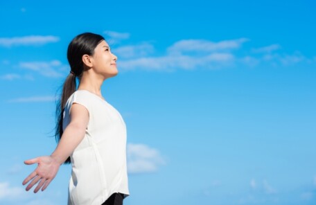 青空の下で大きく両手を広げ、深く呼吸をして微笑む女性。