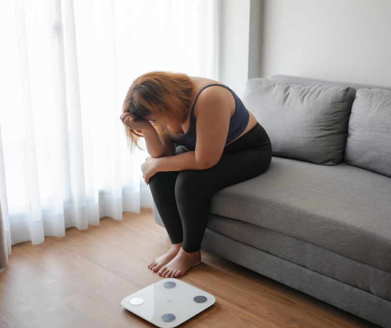 グレーのソファに座り、目の前の体重計を前にして頭を抱え、悩んでいる様子の女性。